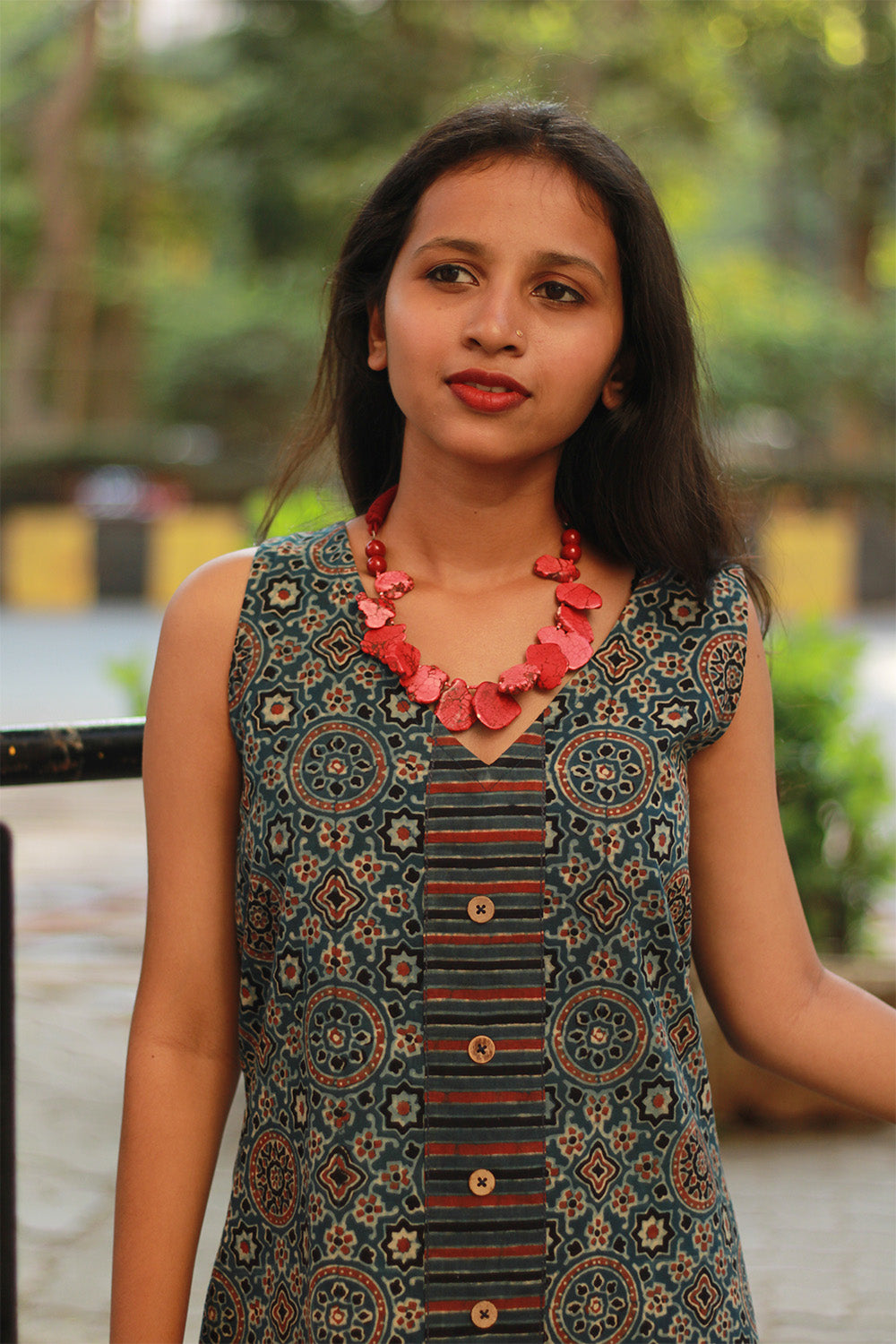 A woman wearing a sleeveless dress with geometric patterns and a red necklace with compressed coral beads.